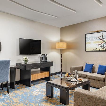Al Ghufran Safwah Hotel Makkah Mekka Buitenkant foto The photo depicts a stylish hotel room or lounge area. It features a comfortable seating arrangement with two sofas and a coffee table in the center. There is a flat-screen television mounted on the wall, alongside a decorative piece of art. A small 
