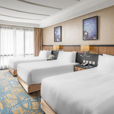 Al Ghufran Safwah Hotel Makkah Mekka Buitenkant foto The photo shows a hotel room featuring three neatly made beds with white linens. The room has a modern and bright design, with large windows allowing natural light to enter. There are two framed artworks on the wall above the beds, contributing to th