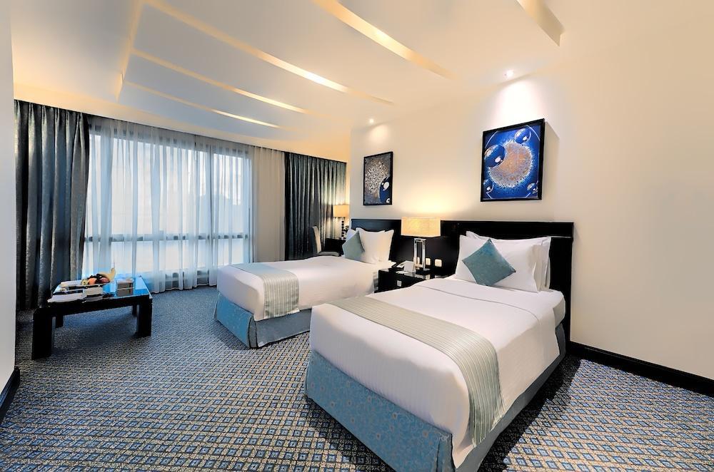 Al Ghufran Safwah Hotel Makkah Mekka Buitenkant foto The photo shows a hotel room with two twin beds. The beds are neatly made with white linens and light blue accents. There are decorative pillows on each bed. A black bedside table with lamps is situated between the beds. On the wall, there are two fr