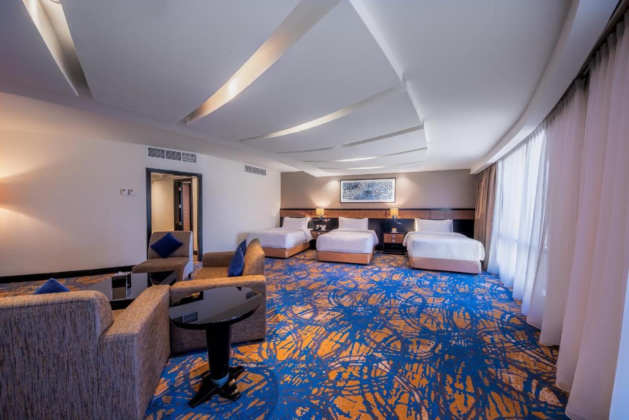 Al Ghufran Safwah Hotel Makkah Mekka Buitenkant foto The photo shows a spacious hotel room with modern decor. It features three white beds arranged neatly, a cozy seating area with a gray sofa and a black coffee table, and large windows letting in natural light. The floor has a vibrant, patterned carpe