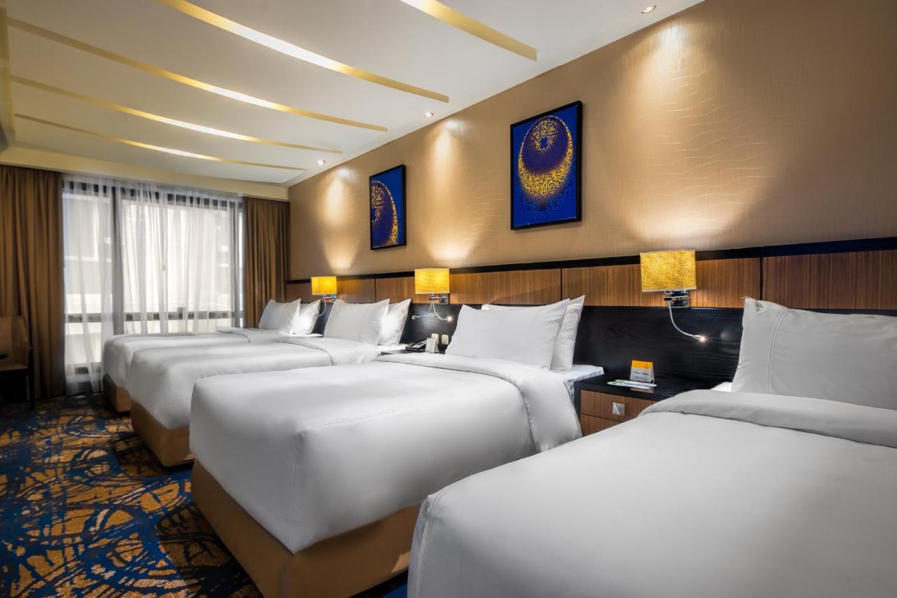 Al Ghufran Safwah Hotel Makkah Mekka Buitenkant foto The image shows a hotel room with four neatly made beds, each covered with white bed linens. The room features warm lighting from bedside lamps and has light brown walls adorned with two decorative blue paintings. The flooring has a patterned carpet,