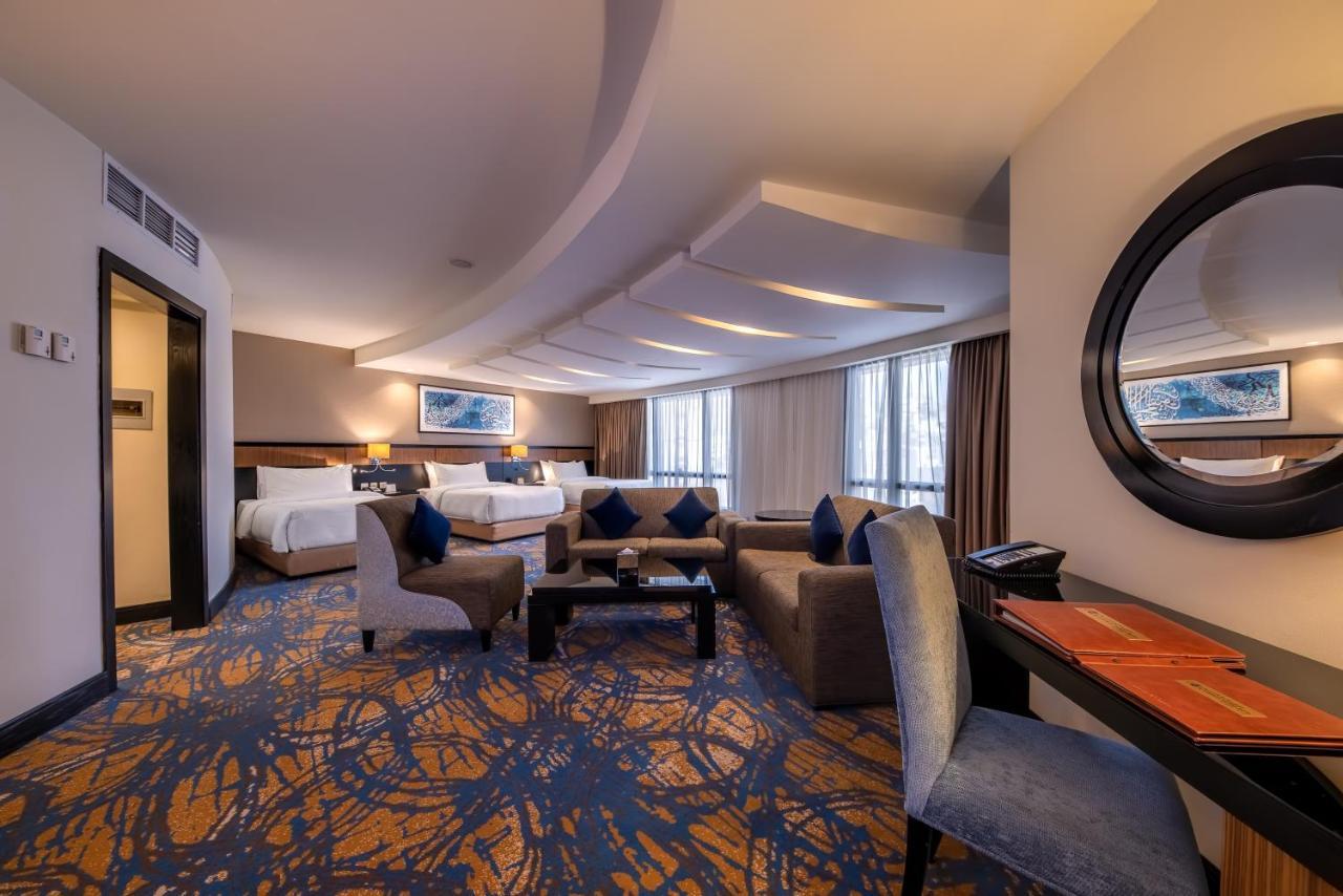 Al Ghufran Safwah Hotel Makkah Mekka Buitenkant foto The photo shows a spacious hotel suite with a modern design. The room features a comfortable seating area with plush chairs and a coffee table, alongside a work desk. There are large windows that let in natural light, and the walls are adorned with a