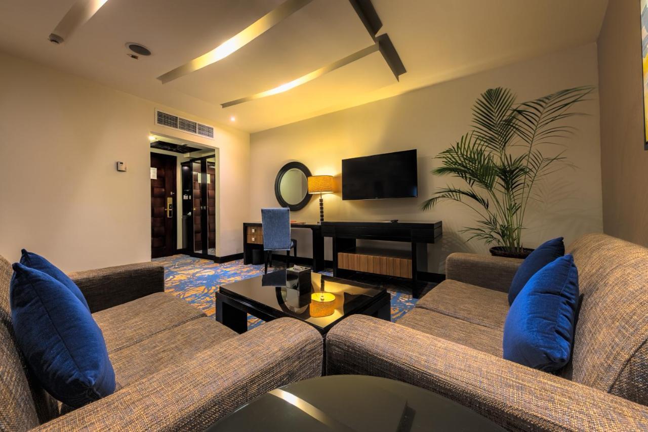 Al Ghufran Safwah Hotel Makkah Mekka Buitenkant foto The photo depicts a modern hotel suite or living area. It features a cozy seating arrangement with a pair of sofas and blue throw pillows. In front of the sofas, there is a glass coffee table with a decorative item on it. 

On the wall opposite the s