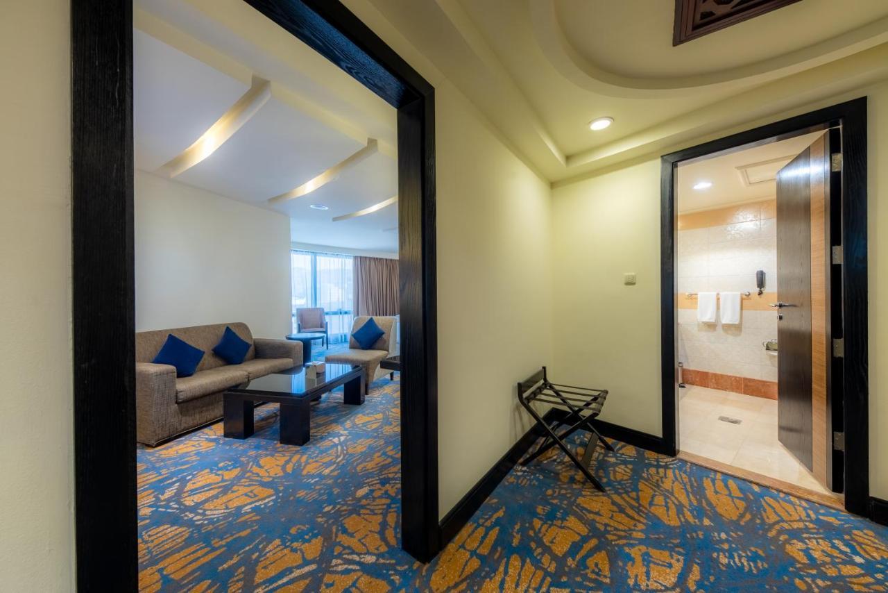 Al Ghufran Safwah Hotel Makkah Mekka Buitenkant foto The photo shows a view of a hotel room entrance. On the left side, there is a seating area with a sofa and a coffee table, decorated with blue cushions. Through a window, natural light illuminates the space. To the right, there is an open door leadin