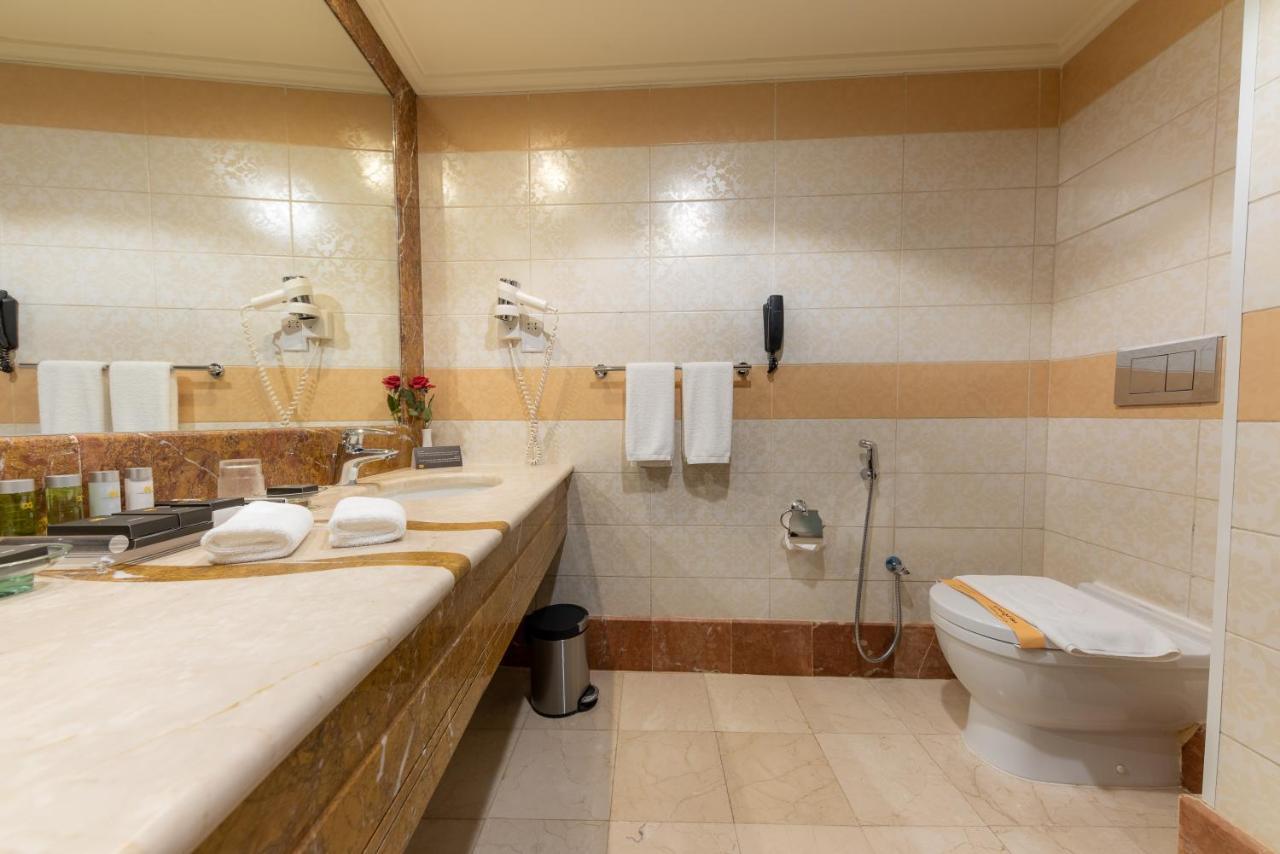 Al Ghufran Safwah Hotel Makkah Mekka Buitenkant foto The photo shows a bathroom that features a large countertop with a sink, which is made of a light-colored stone. There are several toiletries and a small vase with flowers on the countertop. Above the sink, there's a large mirror reflecting the space