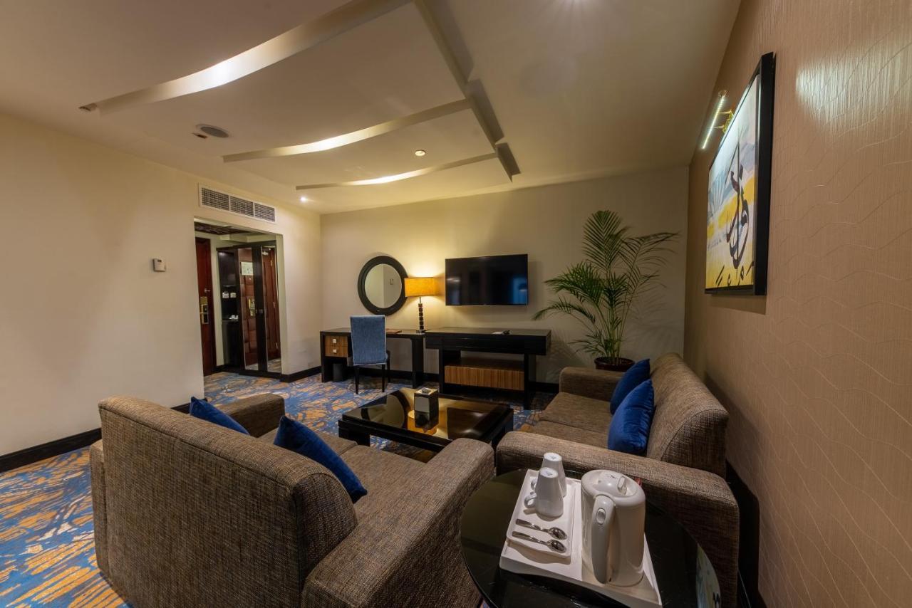 Al Ghufran Safwah Hotel Makkah Mekka Buitenkant foto The photo shows a modern living space that features a cozy seating area. There are gray sofas with blue cushions arranged around a coffee table. A small dining area or workspace with a desk and a chair is visible, along with a flat-screen television 