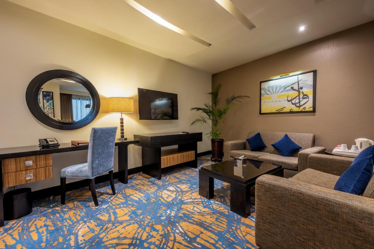 Al Ghufran Safwah Hotel Makkah Mekka Buitenkant foto The photo shows a hotel room or a lounge area. It features a seating arrangement with a light-colored sofa and a matching armchair. There is a coffee table in front, set on a patterned blue and orange carpet. A flat-screen television is mounted on th