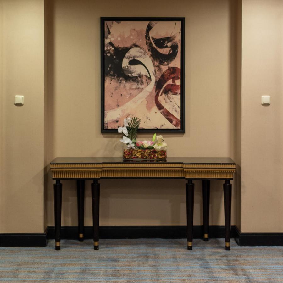 Al Ghufran Safwah Hotel Makkah Mekka Buitenkant foto The photo shows a hallway featuring a decorative wall with a large, abstract painting that includes swirls and shapes in shades of pink and cream. Below the painting, there is a wooden console table with dark legs and a striped design. On the table, 