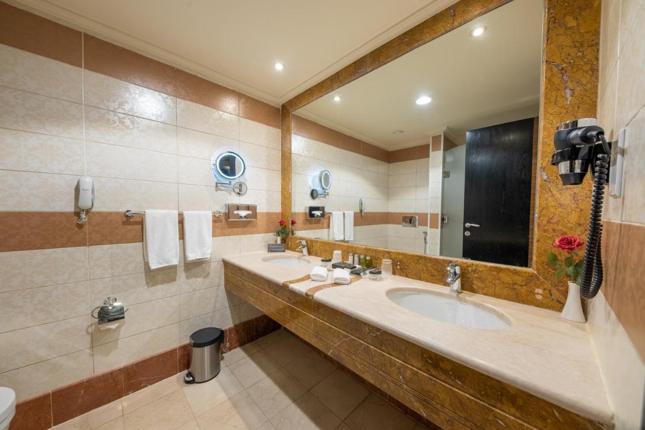 Al Ghufran Safwah Hotel Makkah Mekka Buitenkant foto The photo shows a modern bathroom with a clean and organized design. The walls are tiled with a combination of light and darker colors, and there is a large mirror above a stone countertop. On the countertop, there are toiletries and a small vase wit