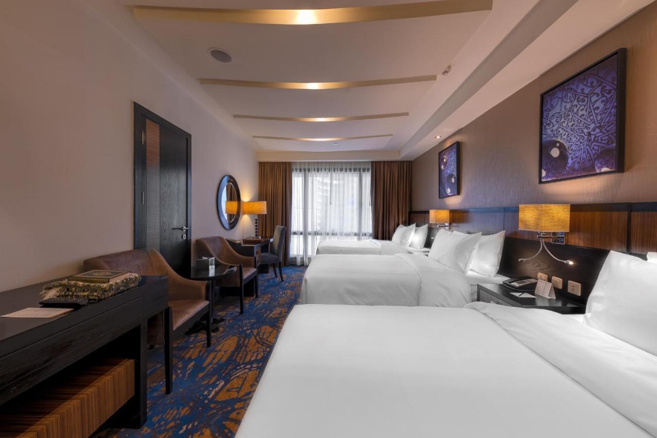 Al Ghufran Safwah Hotel Makkah Mekka Buitenkant foto The photo shows a hotel room featuring two large beds with white bedding. There are two bedside tables with lamps beside each bed. On one side of the room, there is a comfortable chair and a small table, while a work desk can also be seen. The floori