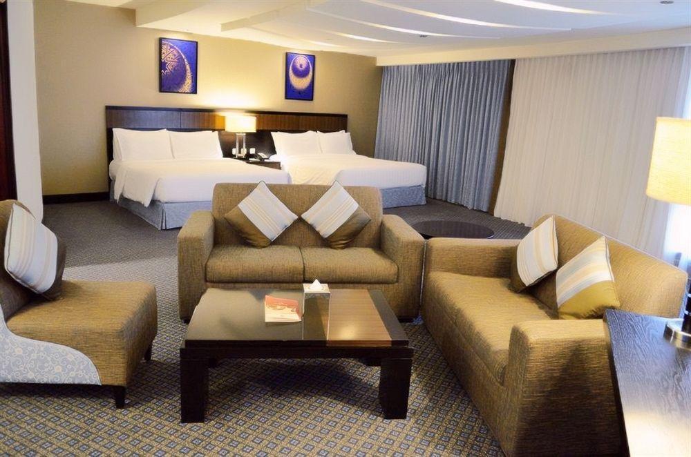 Al Ghufran Safwah Hotel Makkah Mekka Buitenkant foto The photo shows a spacious hotel room featuring a living area and a bedroom. In the foreground, there are two upholstered armchairs and a small coffee table with a book or magazine on it. The couch is L-shaped, accompanied by decorative pillows. In t