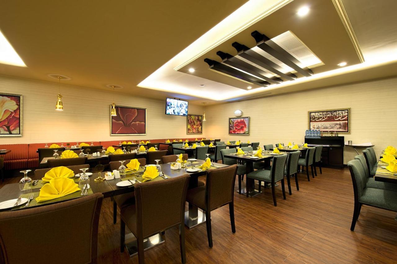 Al Ghufran Safwah Hotel Makkah Mekka Buitenkant foto The image shows the interior of a restaurant. There are several dining tables set for customers, each adorned with yellow napkins and plates. The floors are wooden, and the walls feature paintings or artwork. A flat-screen television is mounted on on
