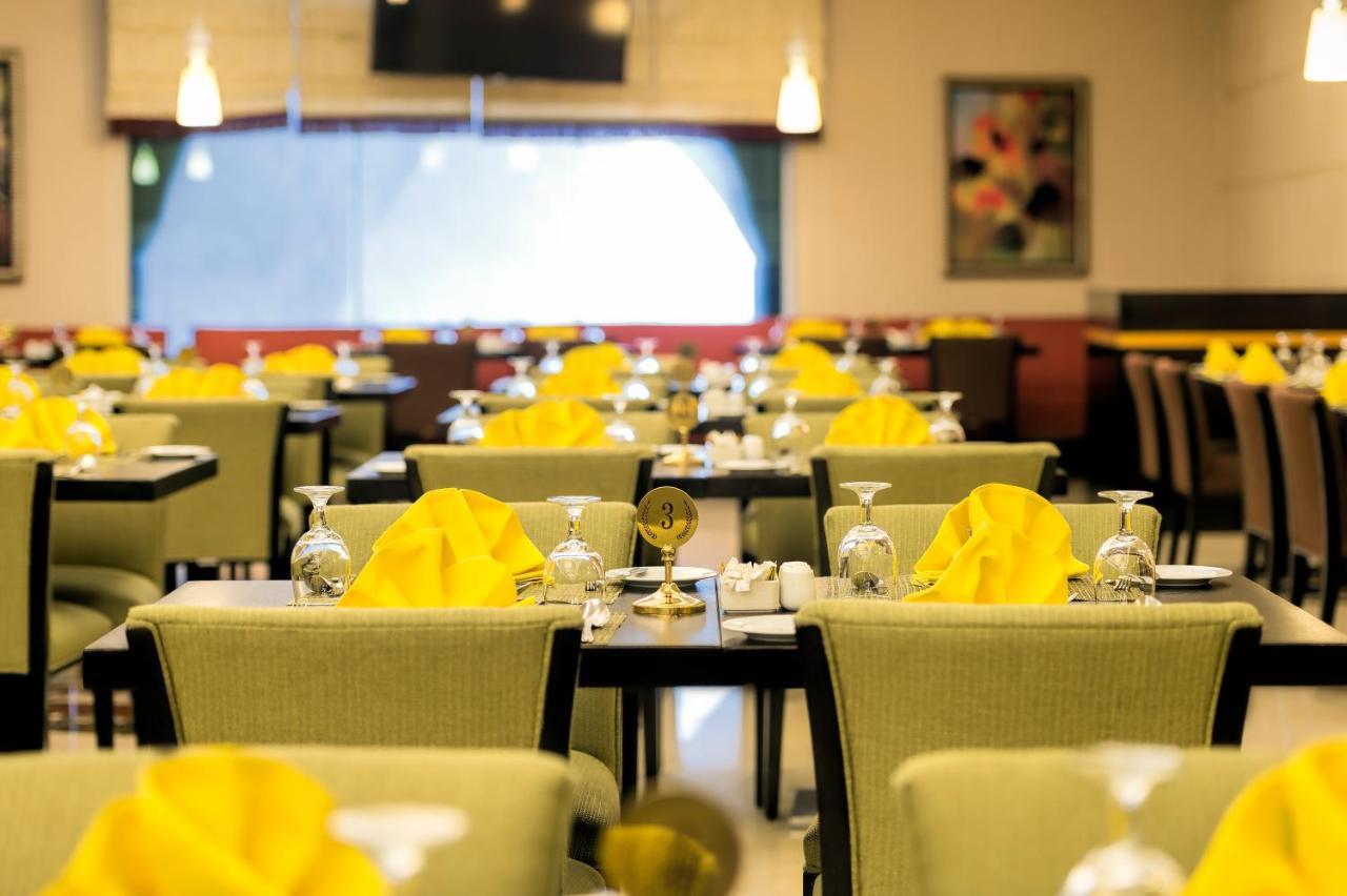 Al Ghufran Safwah Hotel Makkah Mekka Buitenkant foto The photo shows a restaurant interior with several dining tables arranged in a spacious setting. Each table is set with glasses, utensils, and folded yellow napkins. The chairs have a green upholstery, and there's ambient lighting with hanging fixtur