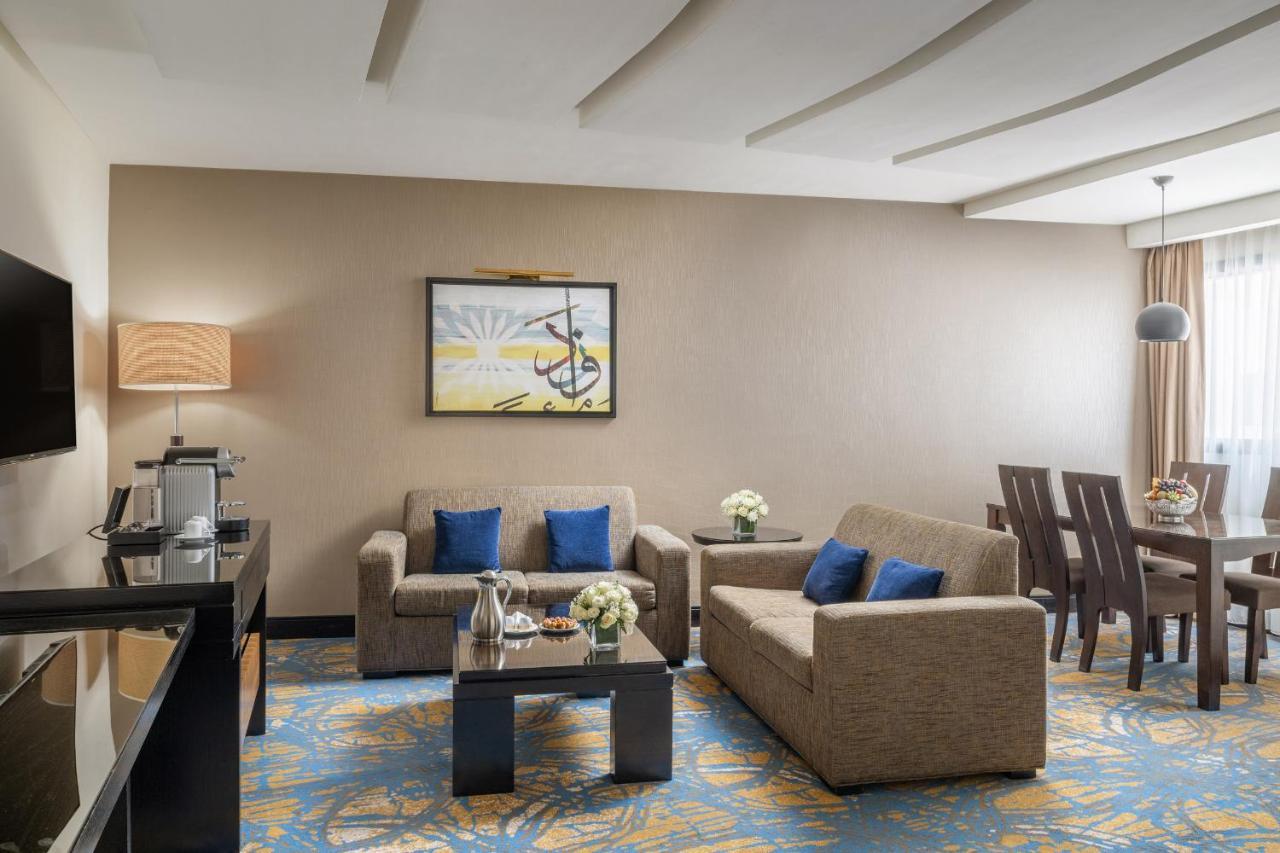 Al Ghufran Safwah Hotel Makkah Mekka Buitenkant foto The photo shows a well-decorated hotel suite or living area. It features a seating arrangement with two beige sofas, adorned with blue cushions. A small coffee table sits in front of the sofas, decorated with flowers. To the side, there's a dining ta