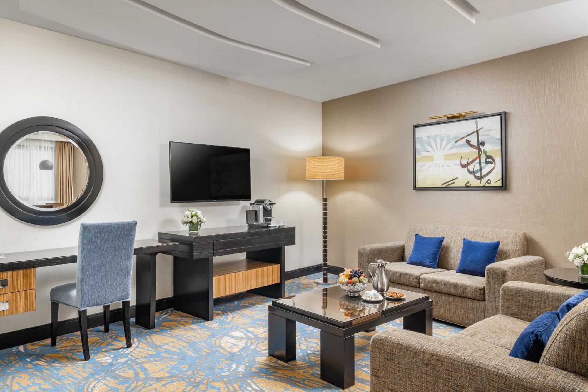 Al Ghufran Safwah Hotel Makkah Mekka Buitenkant foto The photo depicts a stylish hotel room or lounge area. It features a comfortable seating arrangement with two sofas and a coffee table in the center. There is a flat-screen television mounted on the wall, alongside a decorative piece of art. A small 