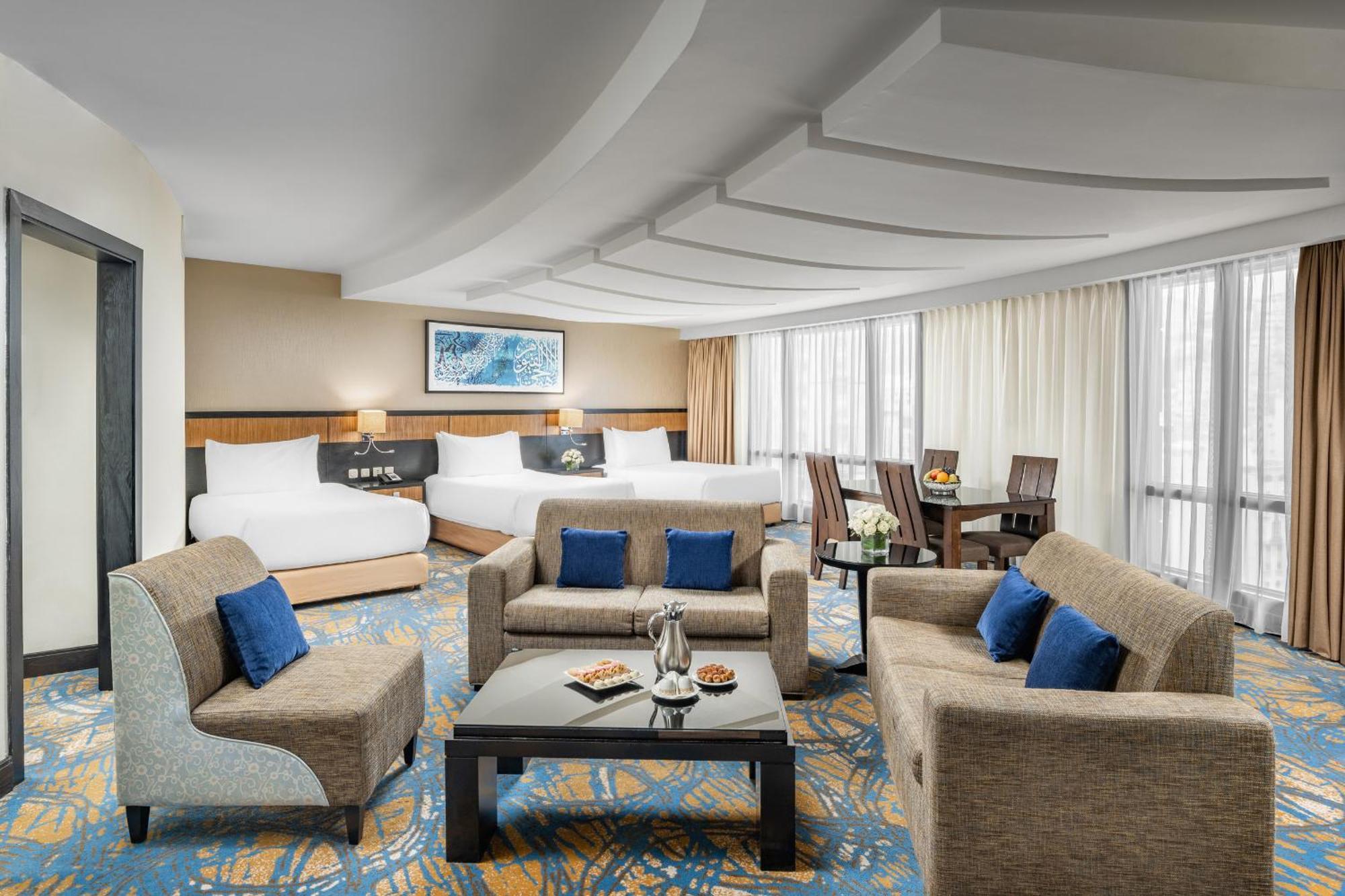 Al Ghufran Safwah Hotel Makkah Mekka Buitenkant foto The photo shows a spacious and elegantly designed hotel room. It features a cozy seating area with a coffee table surrounded by armchairs and a couch, adorned with blue cushions. In the background, there are two beds with white linens, suggesting a c