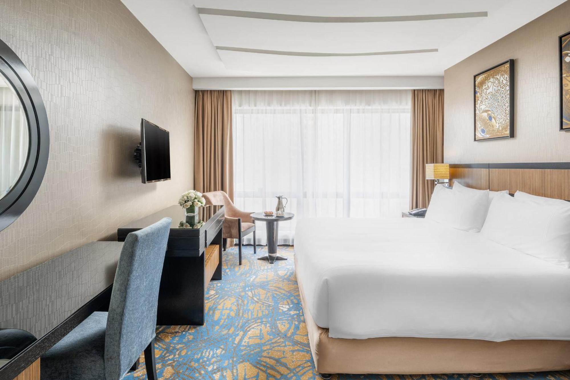 Al Ghufran Safwah Hotel Makkah Mekka Buitenkant foto The photo shows an elegantly designed hotel room. It features a large bed with white linens, a comfy chair, and a small table with two chairs. There is a television mounted on the wall and a decorative piece hanging nearby. The room has large windows
