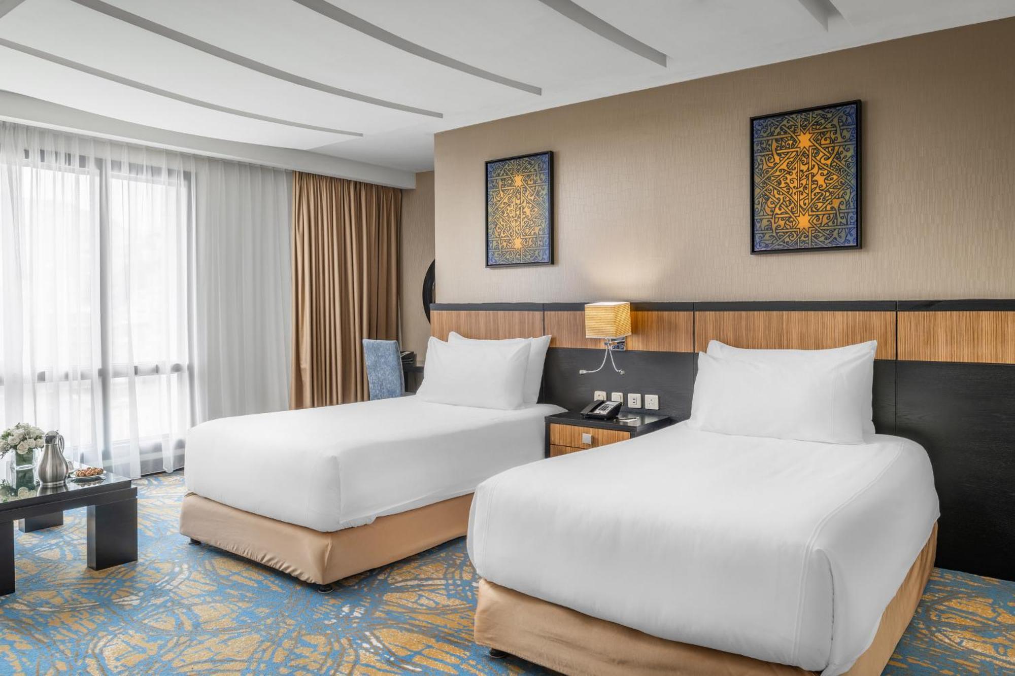 Al Ghufran Safwah Hotel Makkah Mekka Buitenkant foto The photo shows a modern hotel room featuring two white bedding twin beds with soft beige bases. There are decorative paintings on the wall above the beds, and a bedside table with a lamp is positioned between them. The room has large windows covered