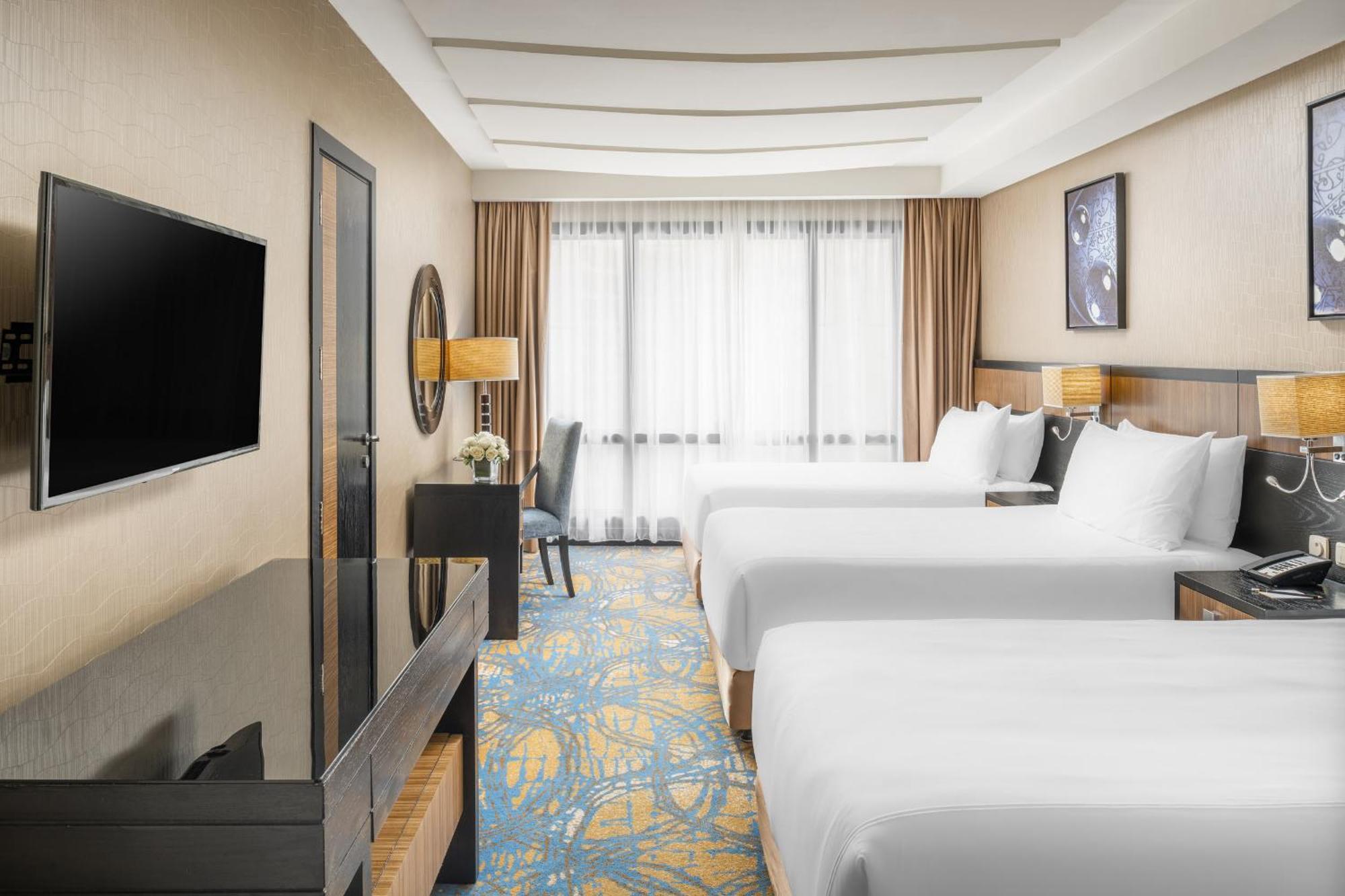 Al Ghufran Safwah Hotel Makkah Mekka Buitenkant foto The photo shows a hotel room that features two large beds with white bed linens. There's a modern, dark wooden furniture design, including a dresser or table and a large TV mounted on the wall. A window with sheer curtains allows natural light to fil