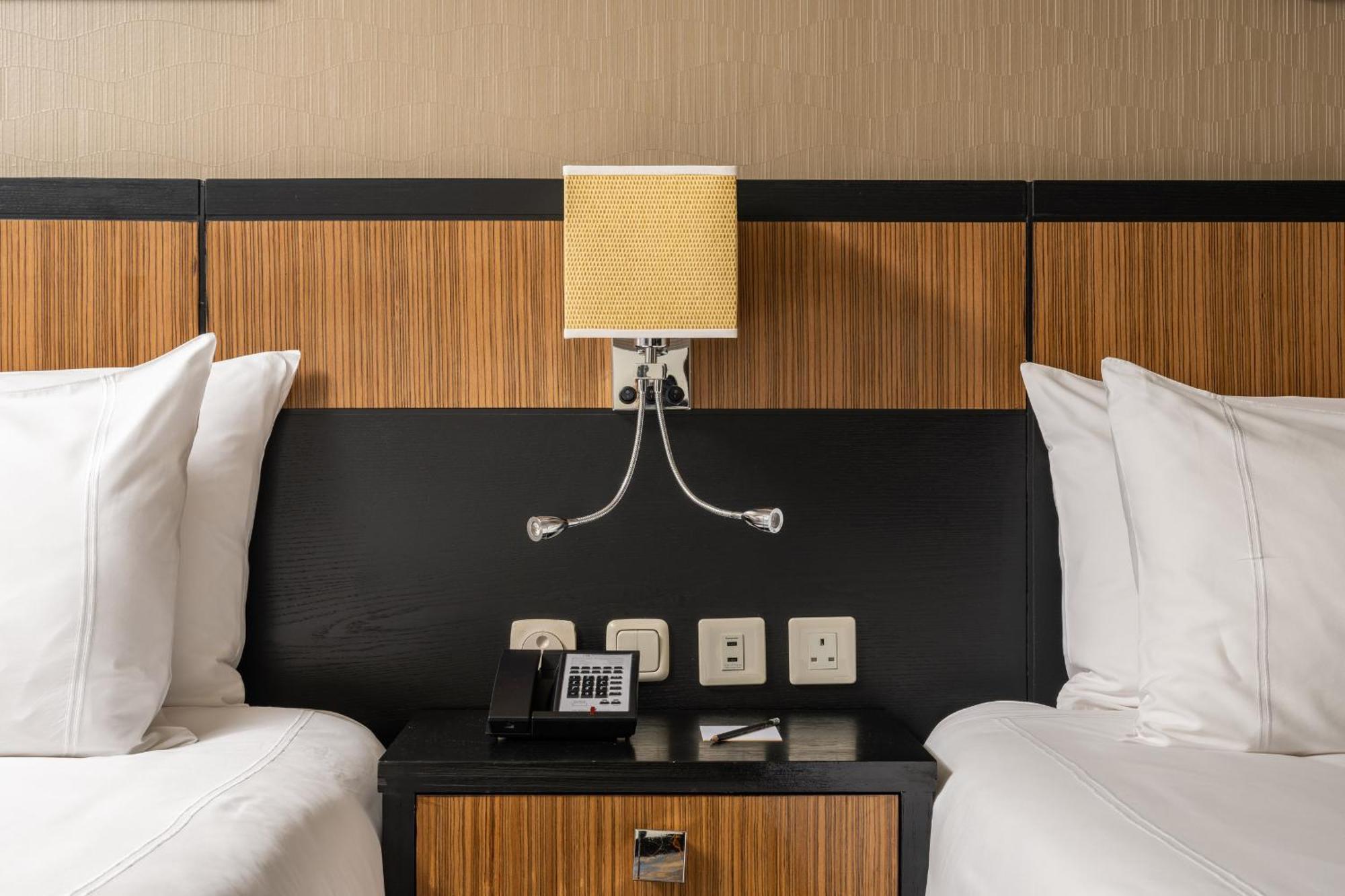 Al Ghufran Safwah Hotel Makkah Mekka Buitenkant foto The photo shows a hotel room with two beds, each adorned with white pillows and bedding. The headboard behind the beds features a combination of wood and dark materials. Above the bedside table, there is a lamp with a yellow shade. The table itself h