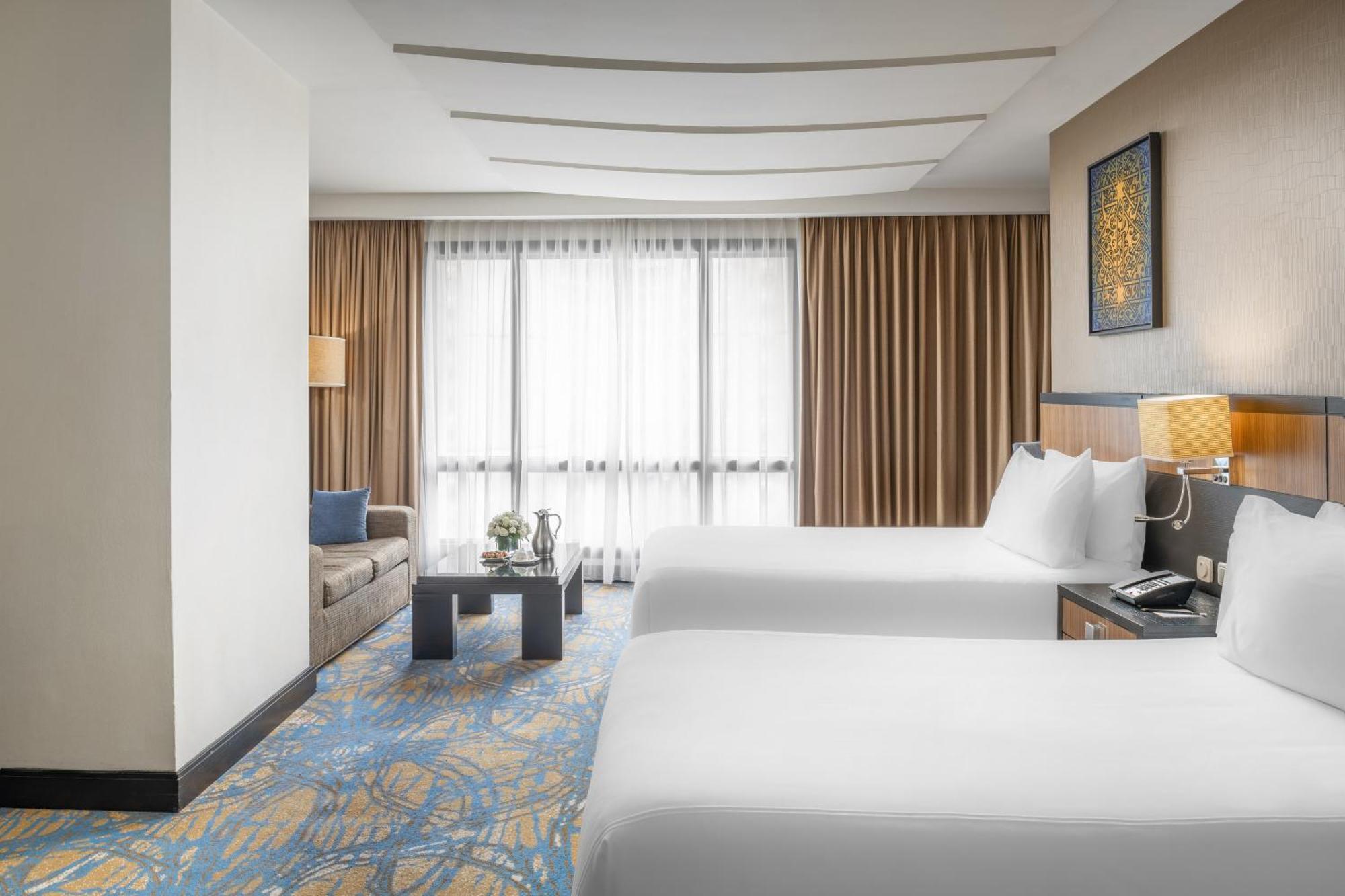 Al Ghufran Safwah Hotel Makkah Mekka Buitenkant foto The photo shows a hotel room with two comfortable beds, dressed in white linens. The decor features a warm color palette, with brown curtains and a stylish, contemporary design. There is a small sofa in the corner beside a coffee table, creating a co