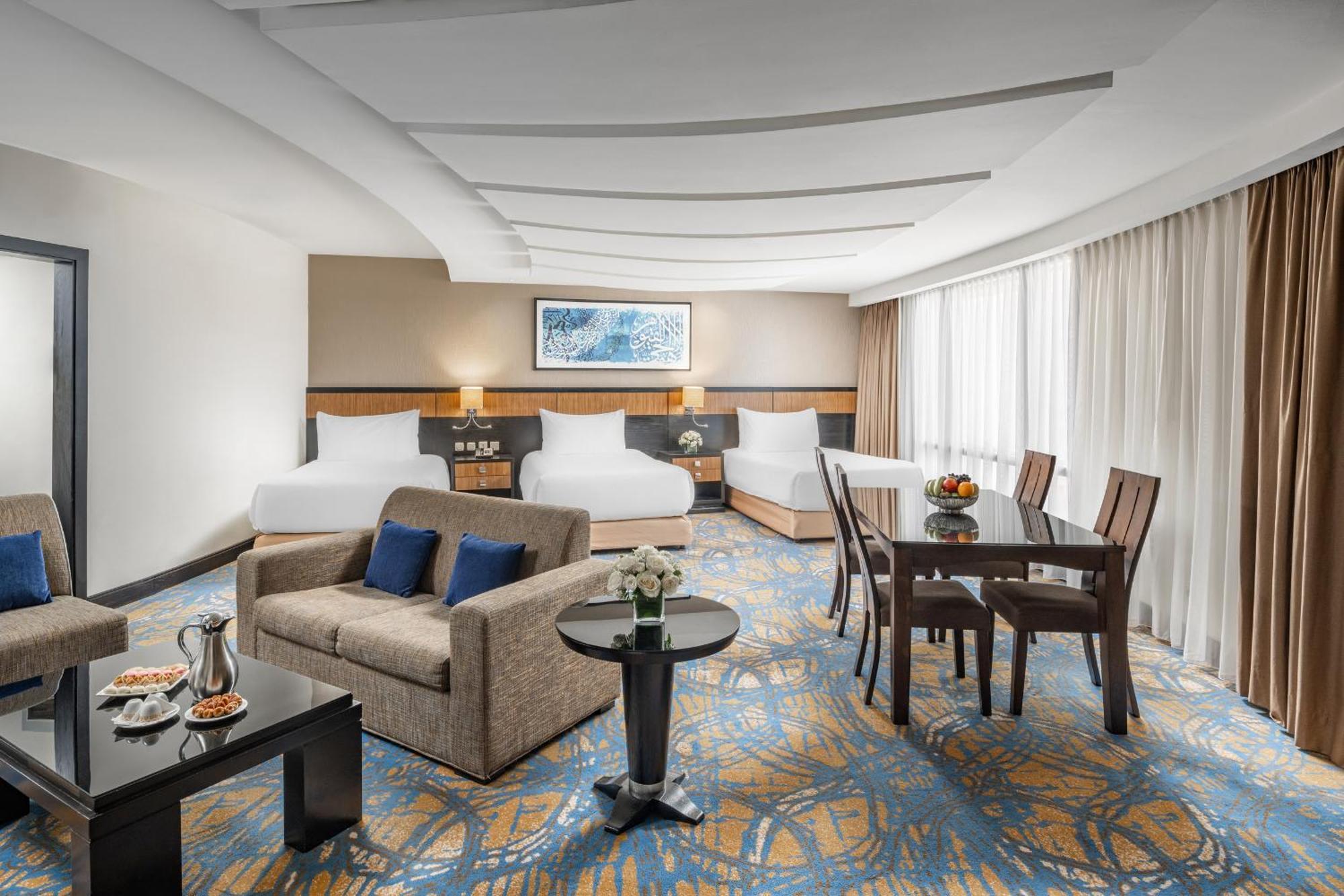 Al Ghufran Safwah Hotel Makkah Mekka Buitenkant foto The photo shows a spacious hotel room designed for comfort. It features two large beds with white bedding, a sitting area with a gray sofa and a coffee table, and a dining area with a wooden table and chairs. There is a decorative vase on the table a