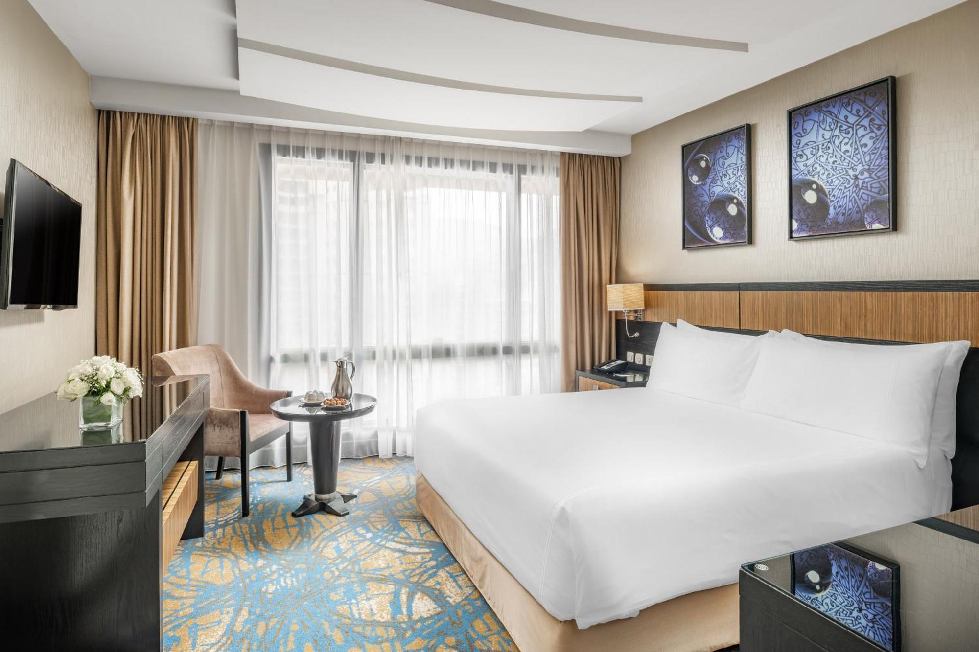 Al Ghufran Safwah Hotel Makkah Mekka Buitenkant foto The image shows a modern hotel room with a spacious layout. It features a large bed with white linens, a bedside table, and stylish wall decor consisting of two framed pictures. There is a comfortable chair and a small round table near the window, wh