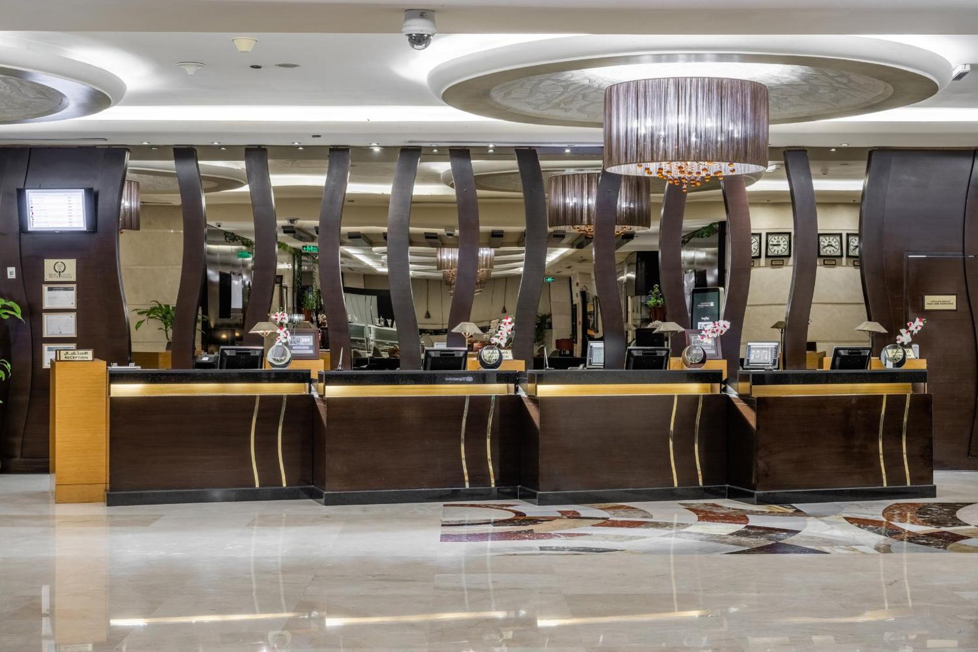 Al Ghufran Safwah Hotel Makkah Mekka Buitenkant foto The photo shows the interior of a hotel lobby, featuring a reception desk with multiple check-in stations. The design includes elegantly curved wooden details and a stylish chandelier overhead. The flooring is polished, enhancing the modern and sophi