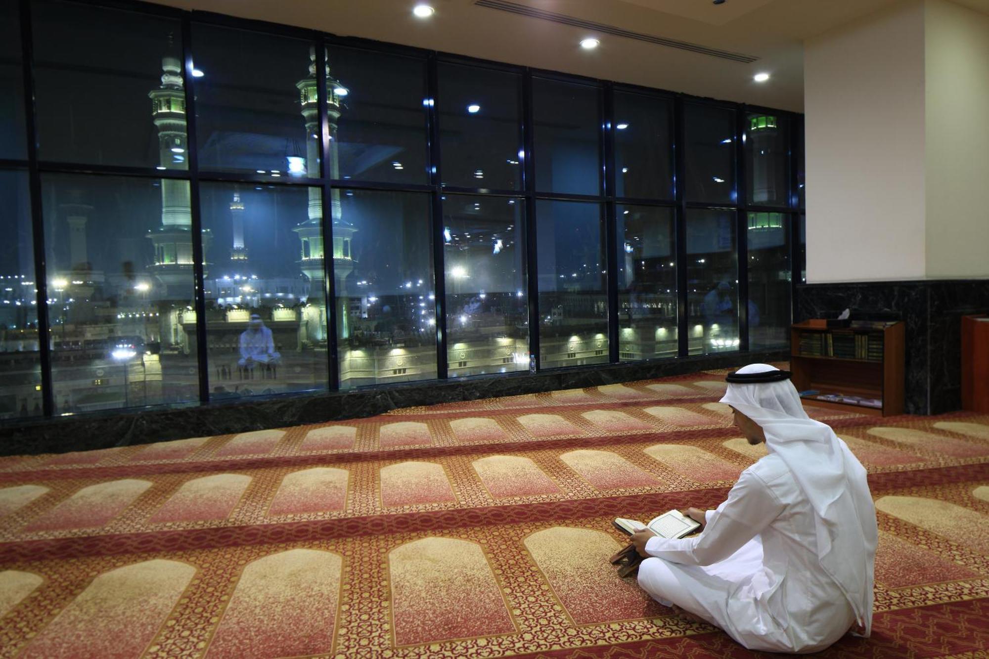 Al Ghufran Safwah Hotel Makkah Mekka Buitenkant foto The photo depicts a person sitting on a carpeted floor, facing away from the camera. They are dressed in traditional attire, likely a white thobe and a headscarf. In the background, large windows reveal a nighttime view of a mosque, with illuminated 
